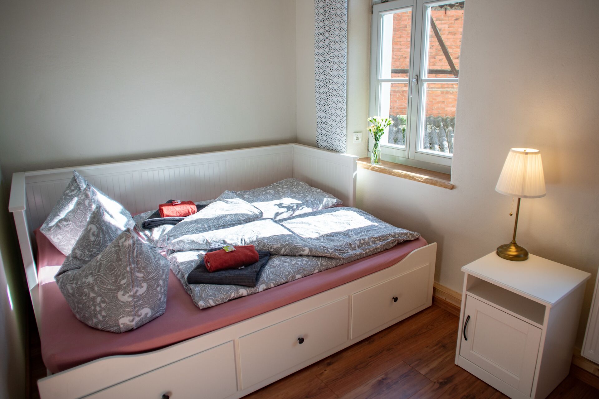 A neatly made bed with patterned bedding, a bedside table with a lamp, and a window with a view of greenery.