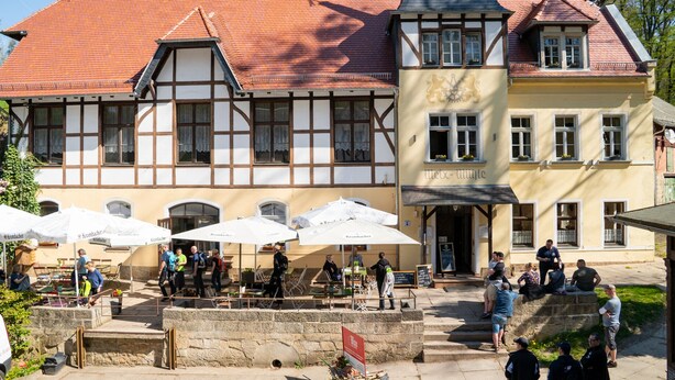 Un bâtiment traditionnel à colombages, avec un toit recouvert de tuiles rouges, un espace extérieur aménagé pour s’asseoir et une enseigne indiquant « Krombacher ».
