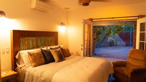 Un dormitorio con una cama, una silla, una mesita de noche y una puerta abierta que da a un patio.