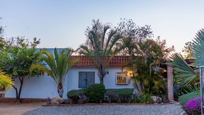 Una casa con techo de tejas rojas, rodeada por un camino de grava, palmeras y diversos arbustos.