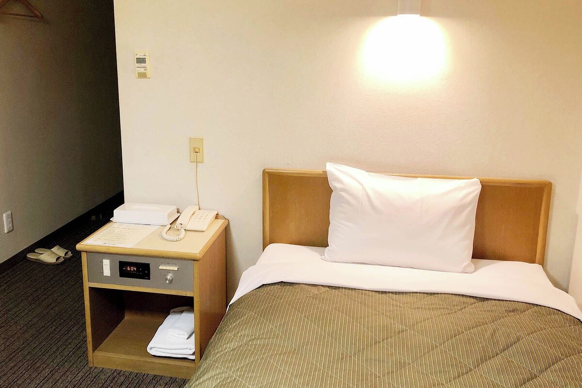 Une chambre d’hôtel avec un lit, une table de chevet avec un téléphone, une lampe et une petite étagère sur laquelle est pliée une serviette.