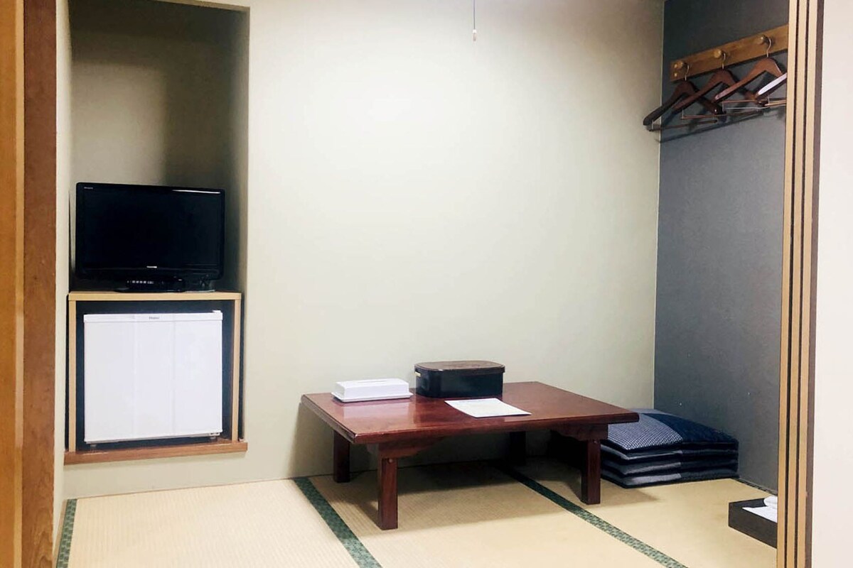 A small room with a television on a shelf, a desk with papers, a small cabinet, and a futon bed.