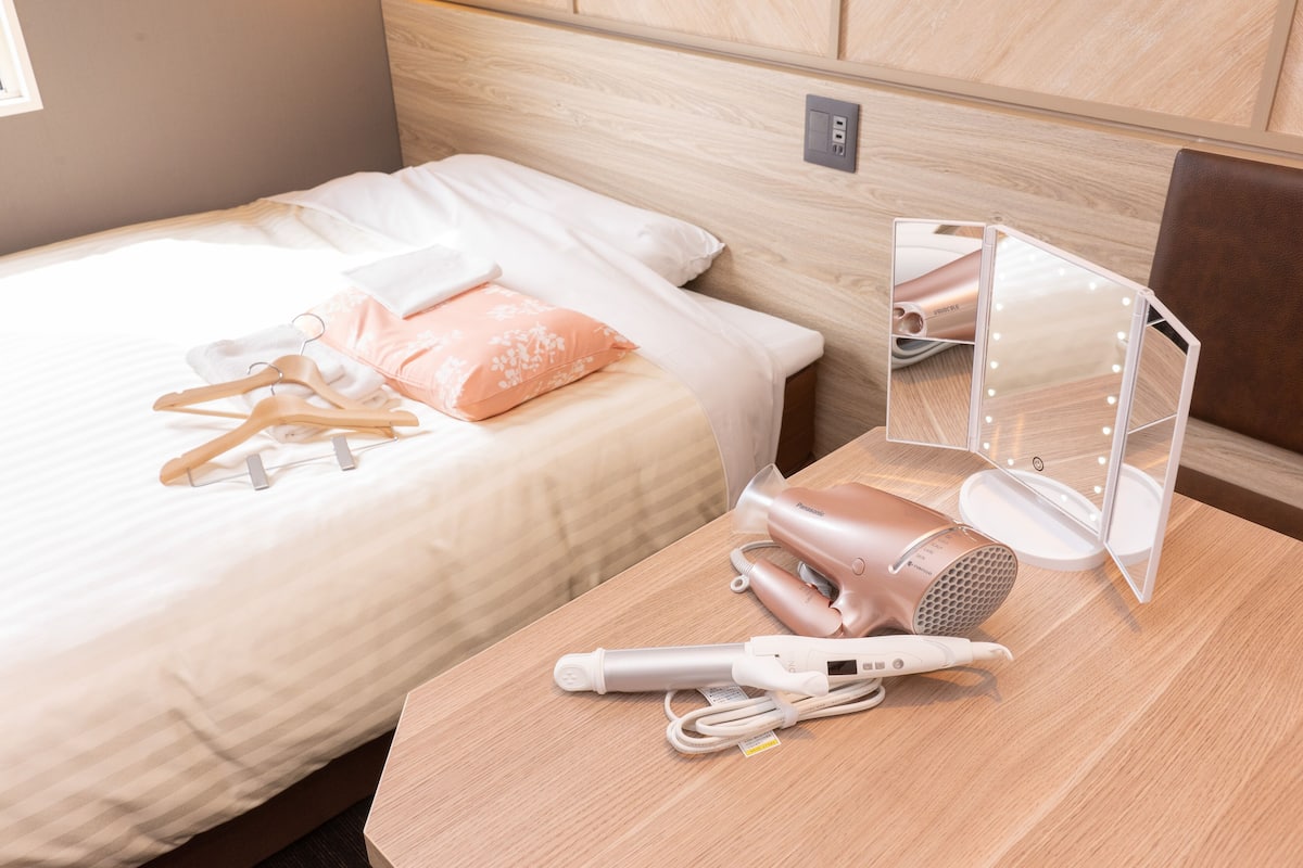 A neatly made bed with a pink pillow, a wooden nightstand with a hairdryer and a mirror, and a wall socket.