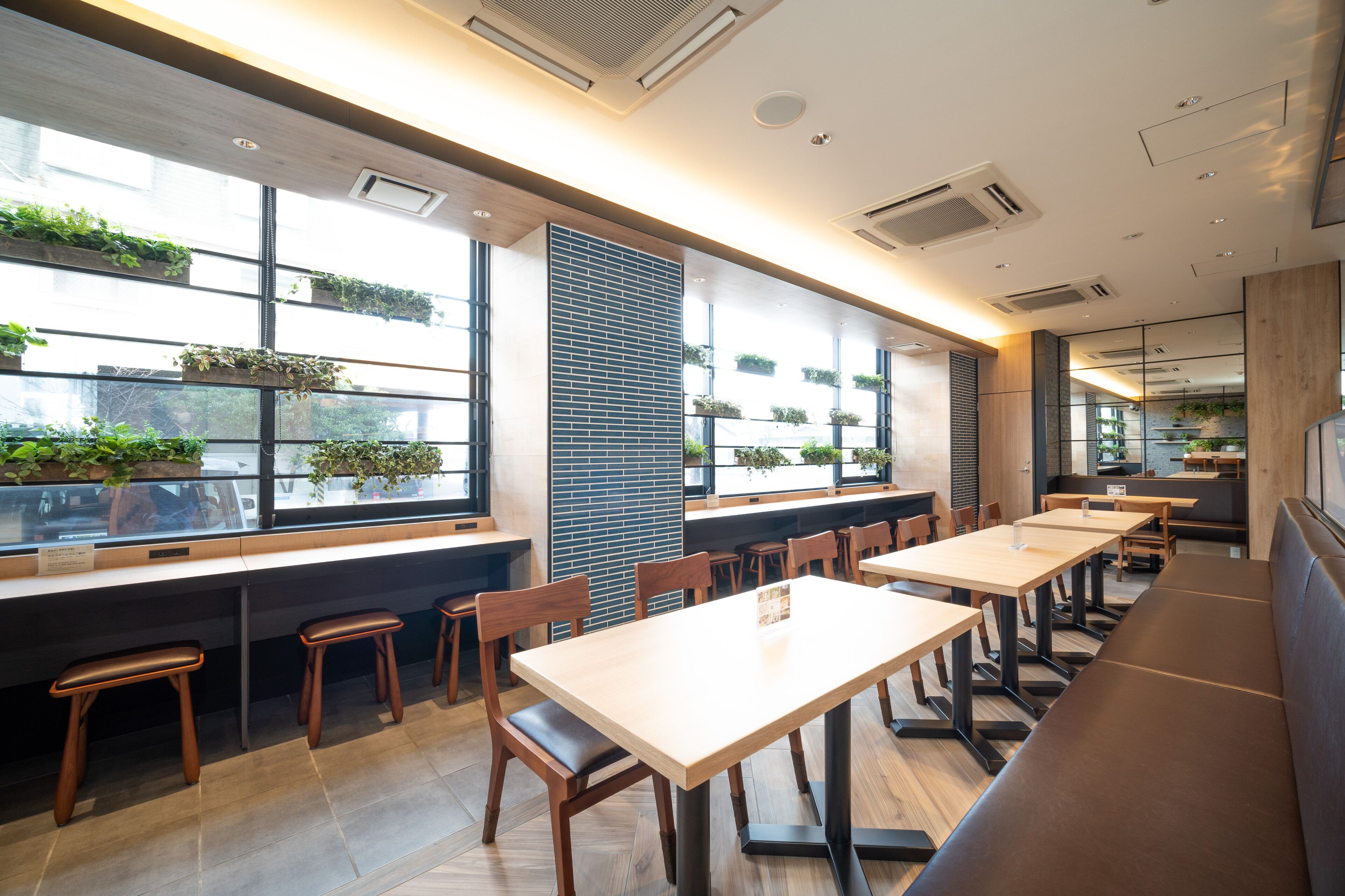 Um restaurante moderno com mesas e bancos compridos, grandes janelas com plantas e um design limpo e contemporâneo.