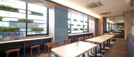 Um restaurante moderno com mesas e bancos compridos, grandes janelas com plantas e um design limpo e contemporâneo.