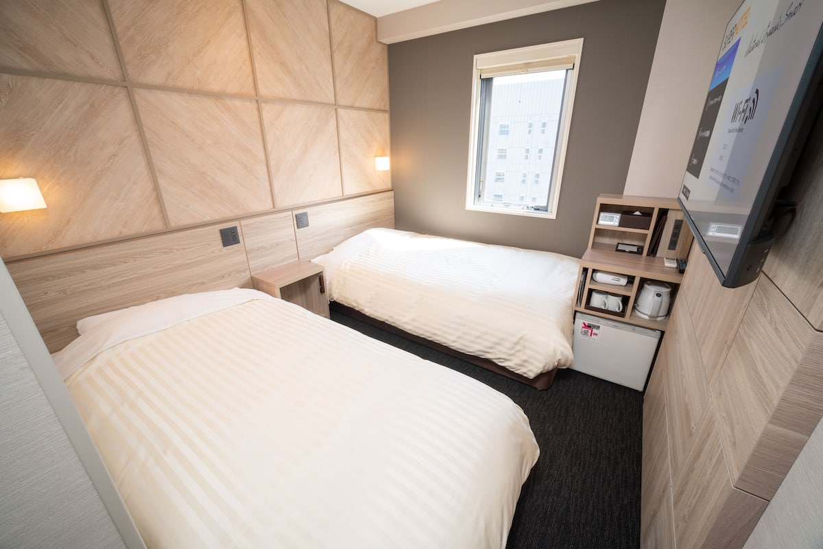 A hotel room with two beds, a small desk, a television, and a window with a view of buildings.