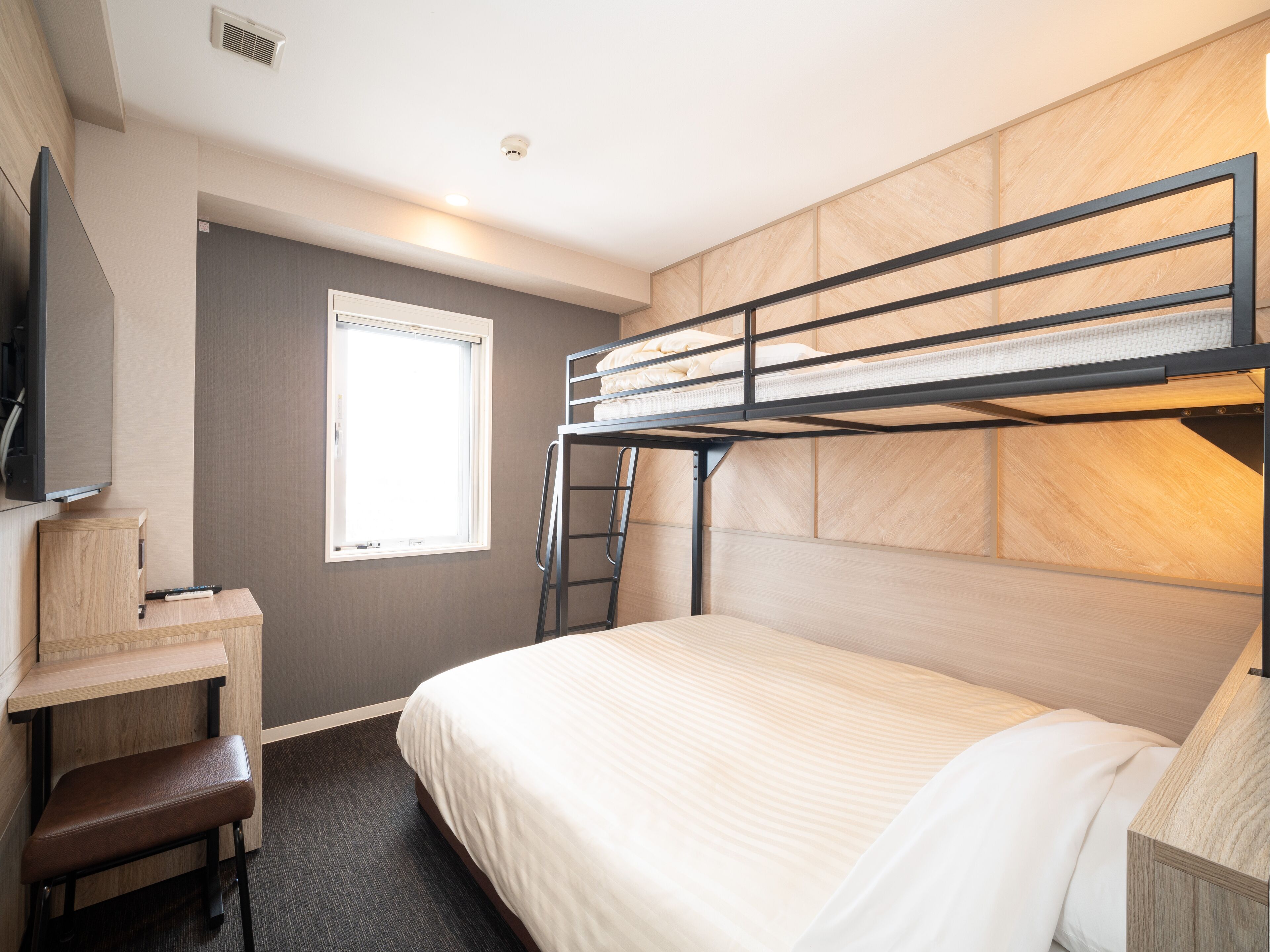A bunk bed room with a desk and chair, a window, and a flat-screen TV mounted on the wall.