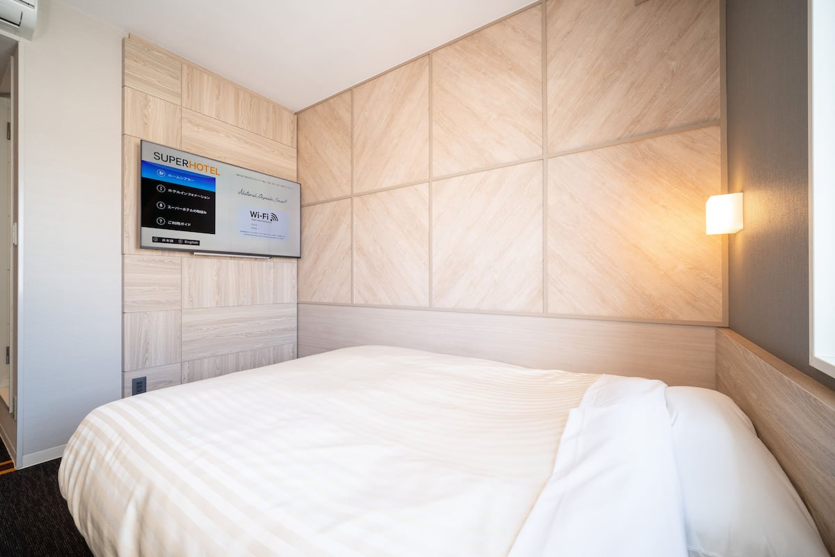 A hotel room with a bed, a wall-mounted TV displaying room information, and a light fixture.