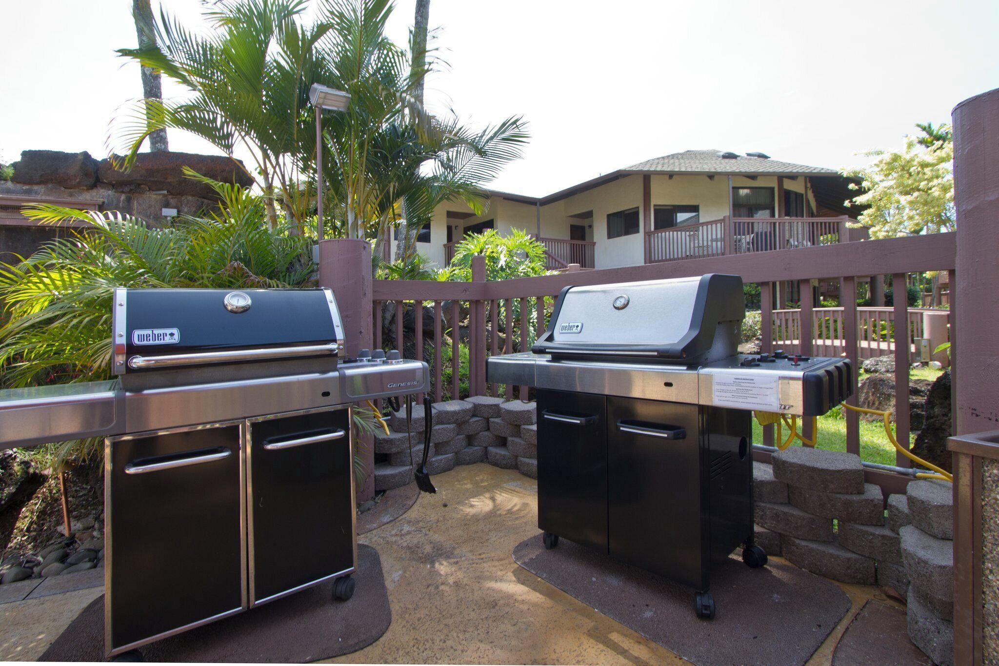 An outdoor barbecue area with two grills, surrounded by a stone wall and palm trees.