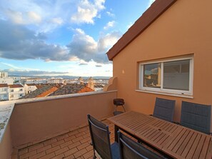 A balcony with a table and chairs, a window, and a view of buildings and the sky.