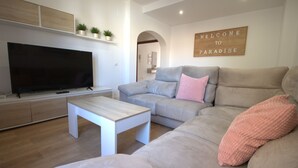 Ein modernes Wohnzimmer mit Fernseher, einem grauen Sofa, einem weißen Couchtisch und einem Schild mit der Aufschrift "WELCOME TO PARADISE". Gesäumt von einem weißen Bücherregal.