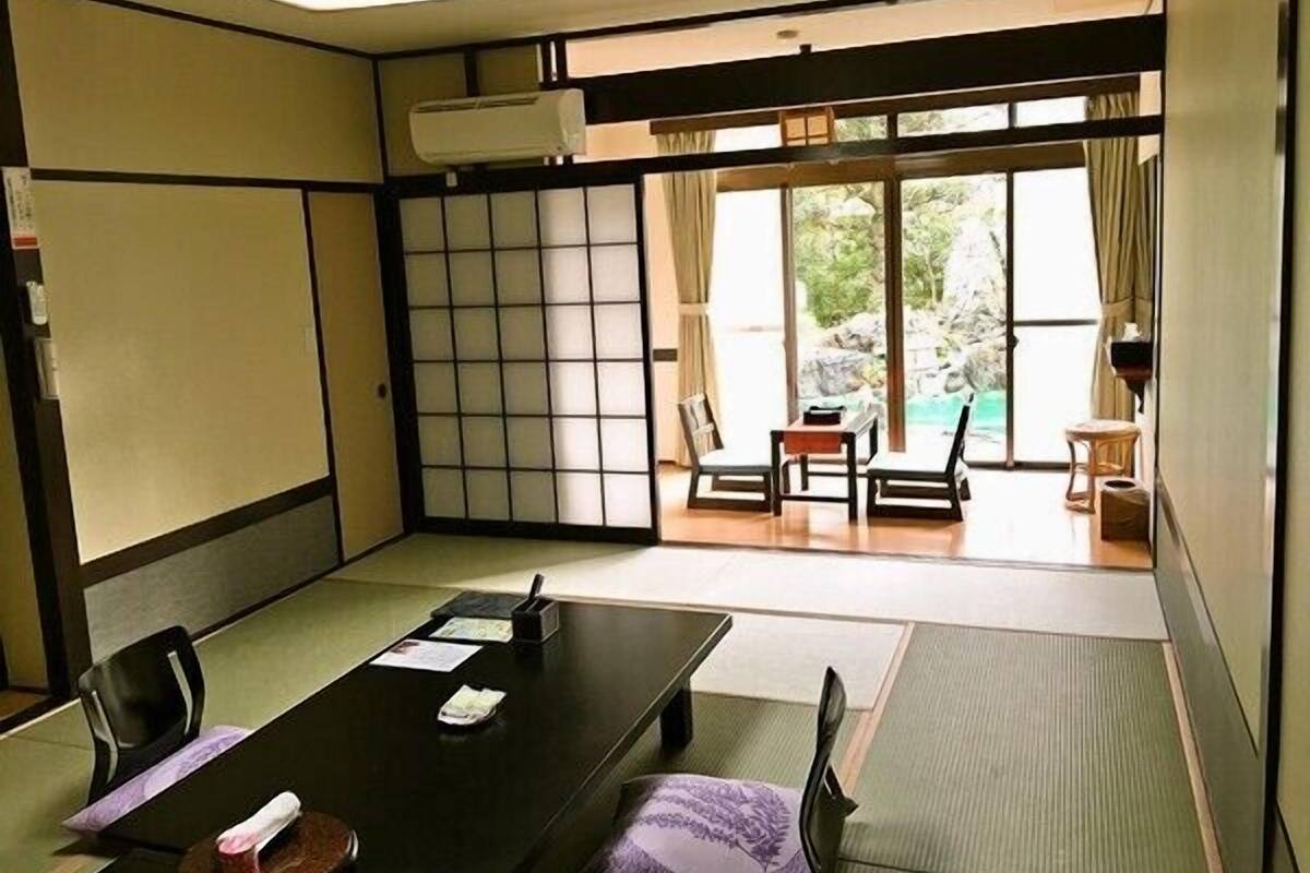 Une pièce de style japonais traditionnel, avec un sol recouvert de tatamis, une table basse entourée de chaises, une porte coulissante donnant sur un balcon avec vue sur un jardin et une piscine.