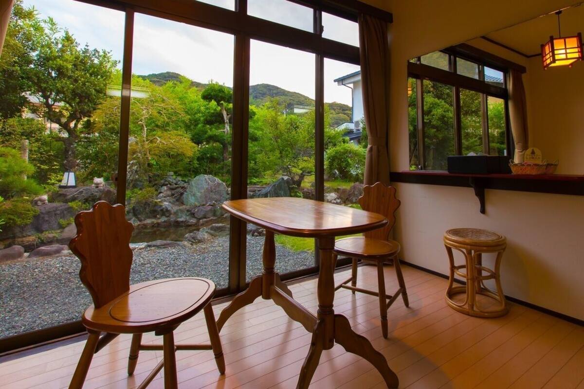 Une pièce avec une table et des chaises en bois, de grandes fenêtres donnant sur un paysage verdoyant et des collines, et un petit tabouret.