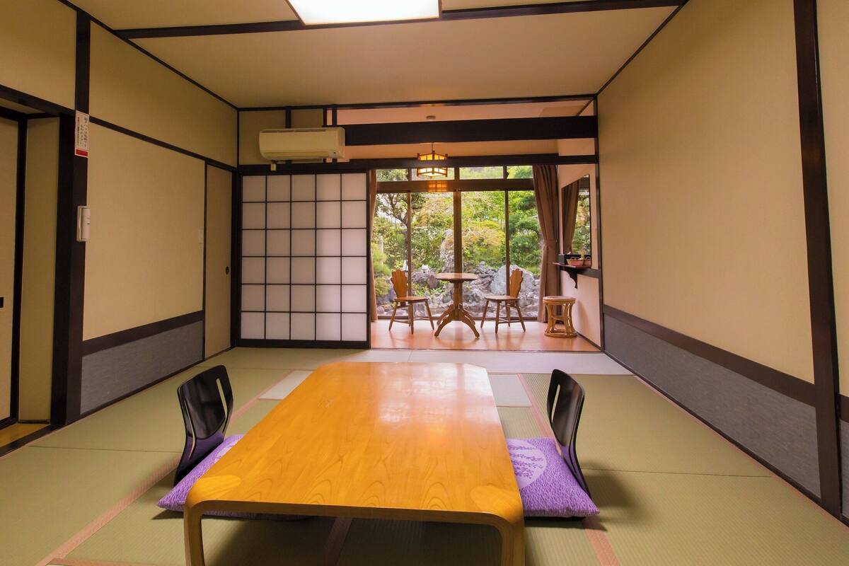 Une pièce de style japonais traditionnel, avec un sol recouvert de tatamis, une table en bois et une vue sur un jardin, accessible par une porte coulissante.