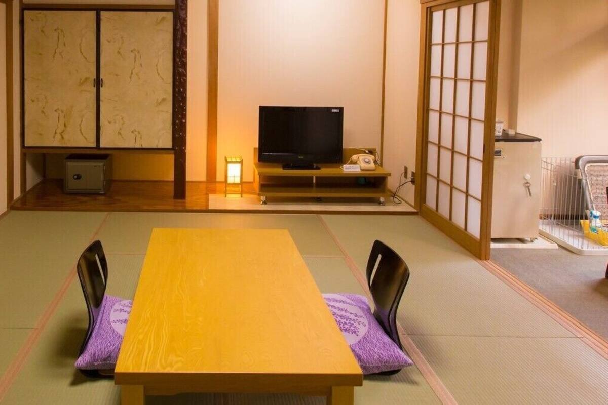 Une pièce de style japonais traditionnel, avec un sol recouvert de tatamis, une table basse entourée de chaises, un téléviseur et une porte coulissante.