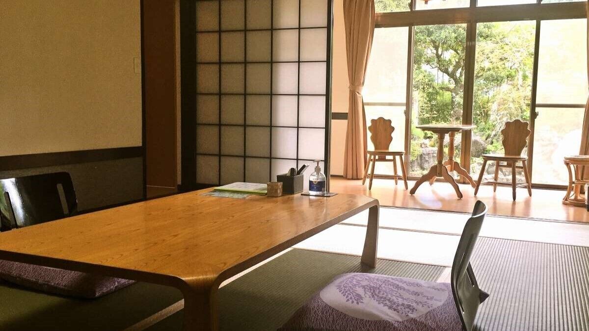A traditional Japanese-style room with tatami flooring, a wooden table, and a view of greenery through large windows.