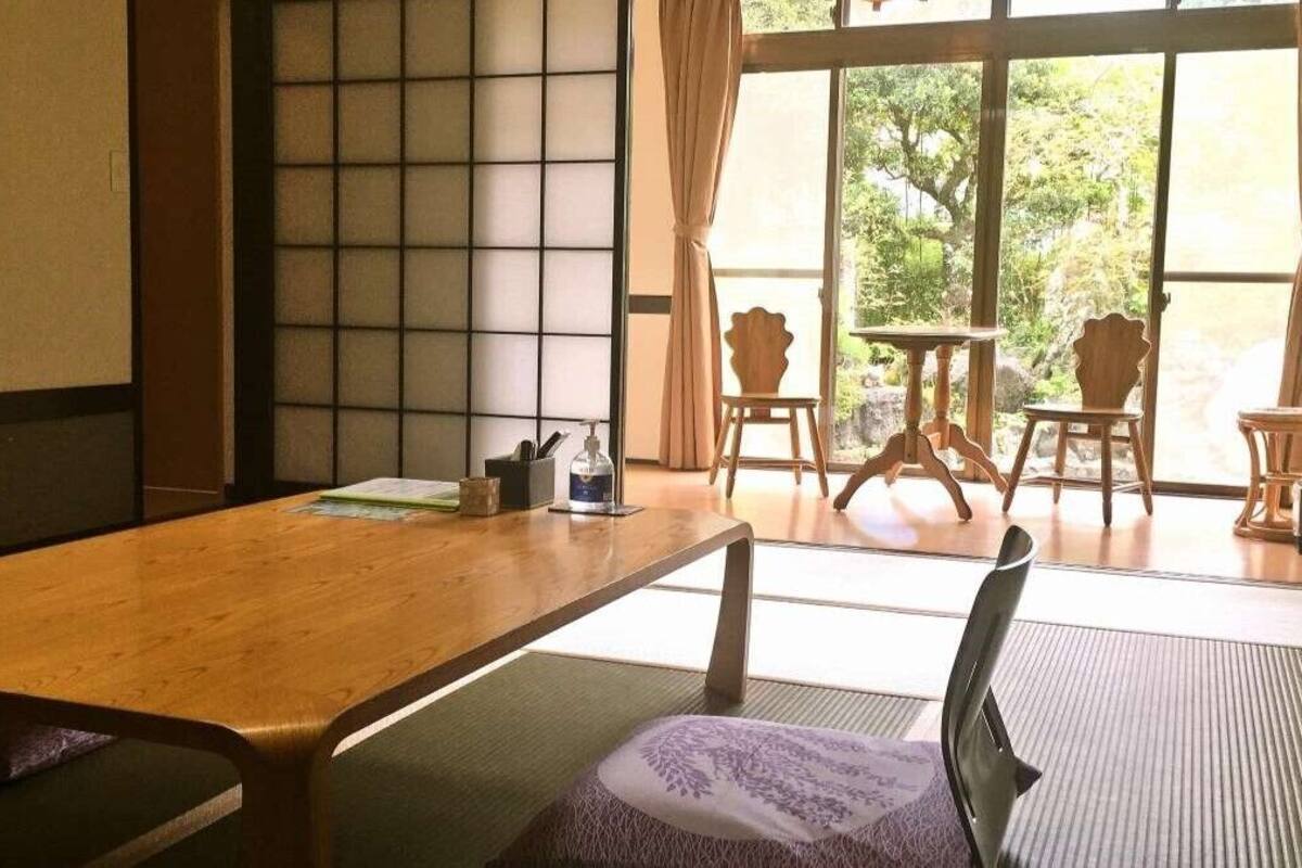 Une pièce de style japonais traditionnel, avec un sol recouvert de tatamis, une table en bois et une vue sur la verdure grâce à de grandes fenêtres.