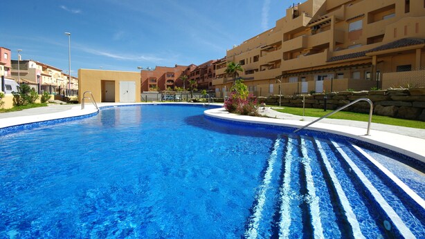 Une piscine avec un bassin aux eaux d’un bleu limpide, entourée d’immeubles à plusieurs étages dotés de balcons et d’un petit jardin agrémenté de fleurs roses.