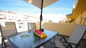 Une table avec un plateau en verre, deux chaises, une bouteille de vin, une pastèque et quelques citrons, le tout sur un balcon d’où l’on peut admirer des immeubles.