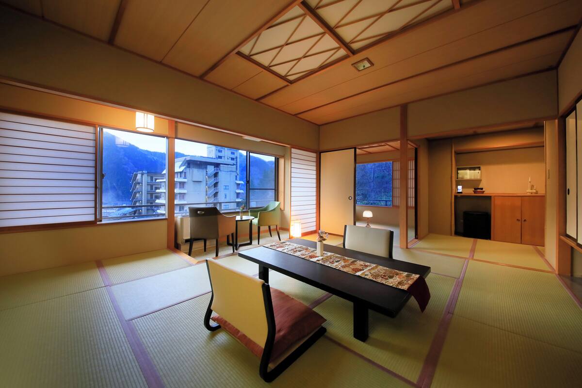 Una habitación japonesa tradicional con suelo de tatami, una mesa baja con un mantel individual, una silla acolchada y puertas correderas que dan a un balcón con vistas a edificios y montañas.