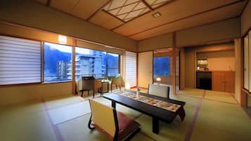A traditional Japanese room with tatami flooring, a low table with a placemat, a cushioned chair, and sliding doors leading to a balcony with a view of buildings and mountains.