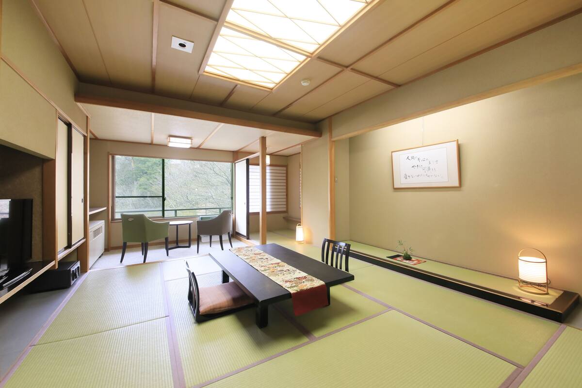 Una habitación tradicional japonesa con suelo de tatami, una mesa baja, sillas, un televisor y una ventana con vistas a la vegetación.