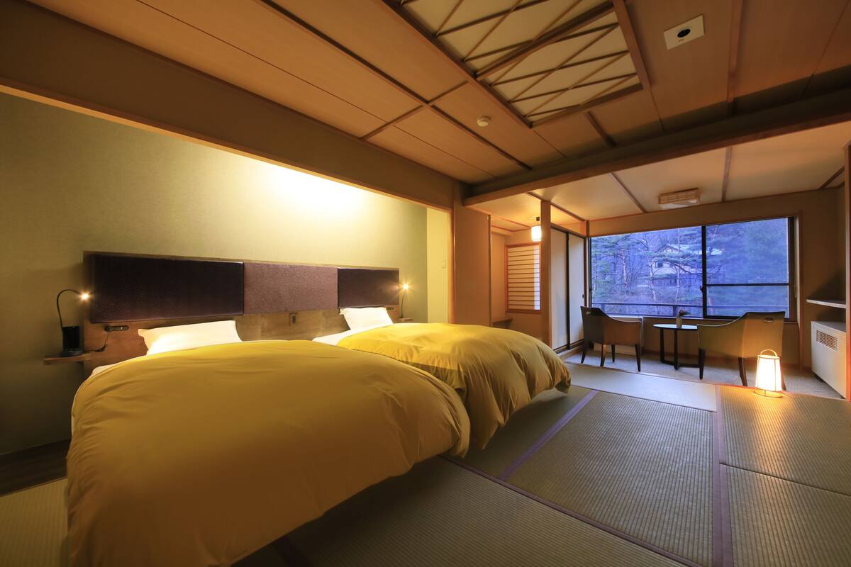 Habitación de hotel con dos camas, una zona de estar con una mesa y sillas, y vistas a árboles por la ventana.