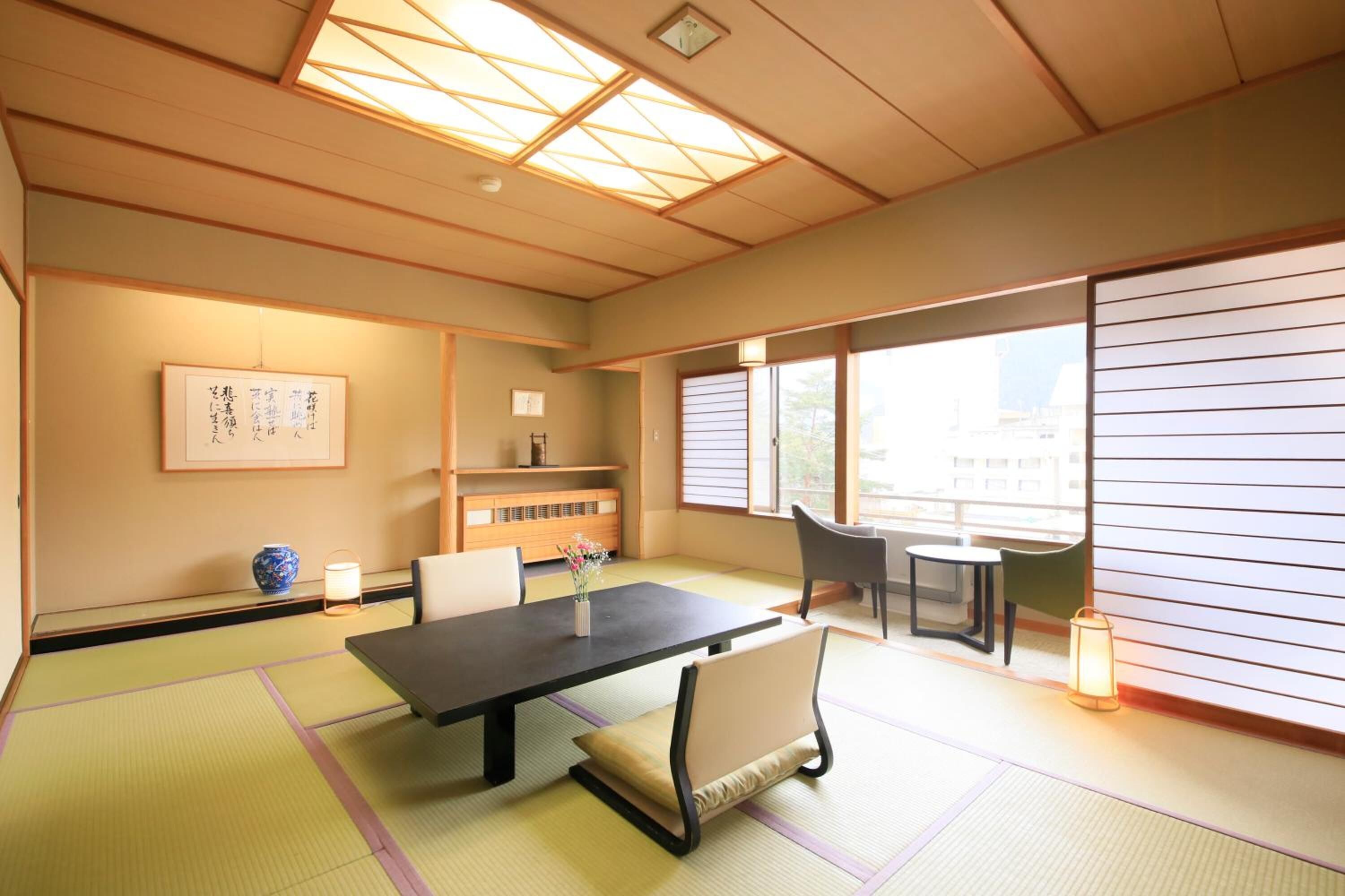 Una habitación japonesa tradicional con suelo de tatami, una mesa baja, sillas, una mesita con una lámpara y un gran ventanal con puertas correderas.