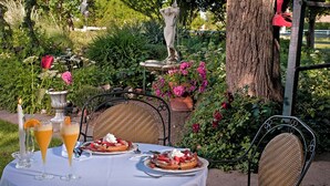 An outdoor dining area with a table set for two, surrounded by lush greenery and a statue.