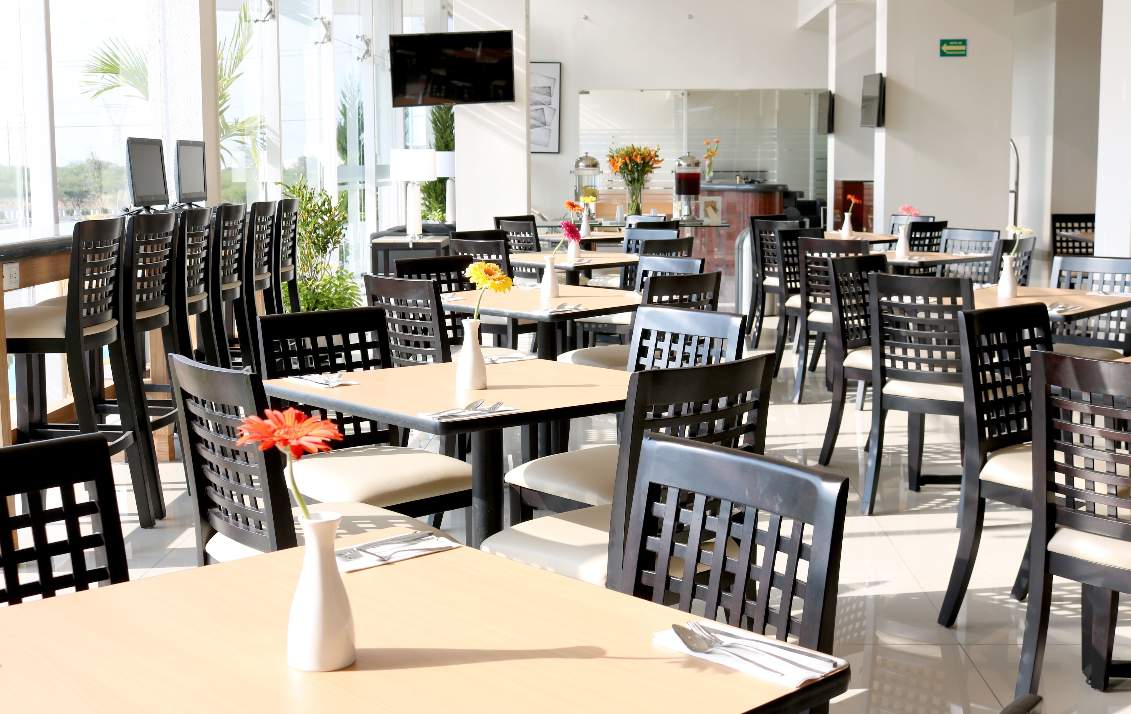 A spacious restaurant with multiple tables and chairs, large windows, and a television mounted on the wall.