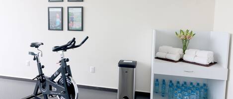 Zona de gimnasio con una bicicleta estática, un cesto de basura, un reloj y un estante con toallas y botellas de agua.
