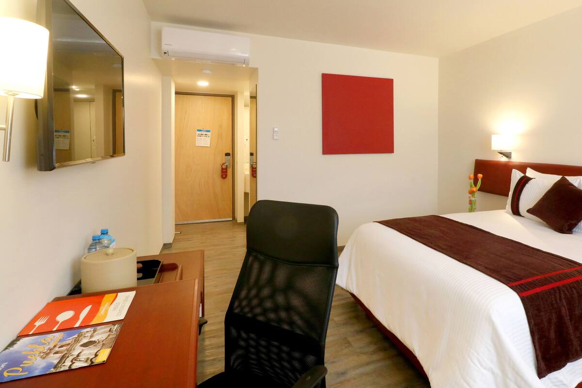 A hotel room with a bed, a desk, a chair, a mirror, and a red wall decoration.