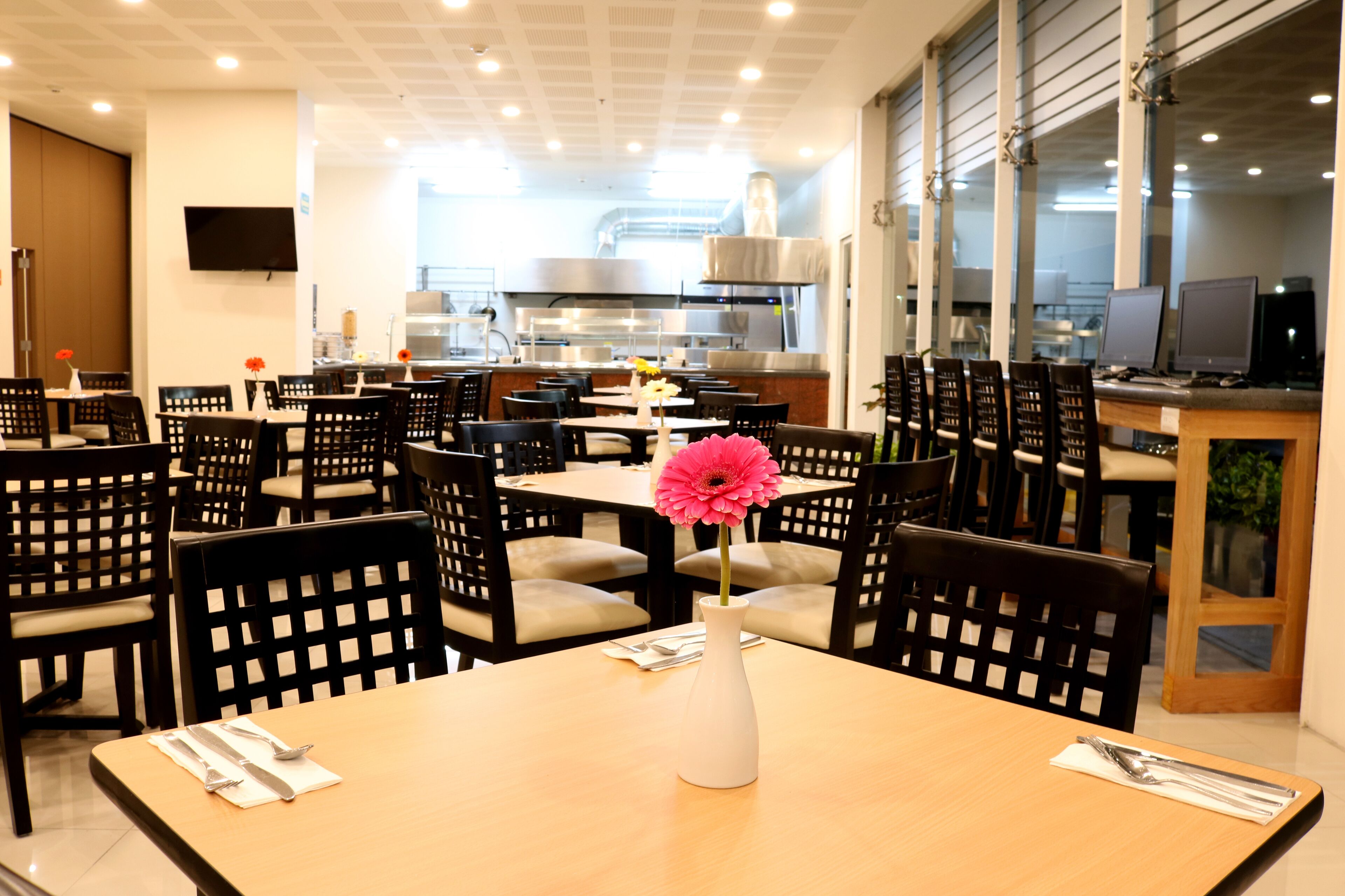 Un restaurante con varias mesas y sillas, un florero blanco con una flor rosa, y una zona de cocina al fondo.