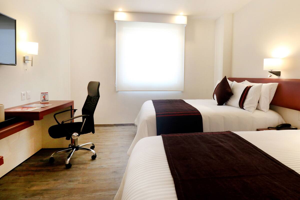 A hotel room with two beds, a desk with a chair, a TV, and a window.