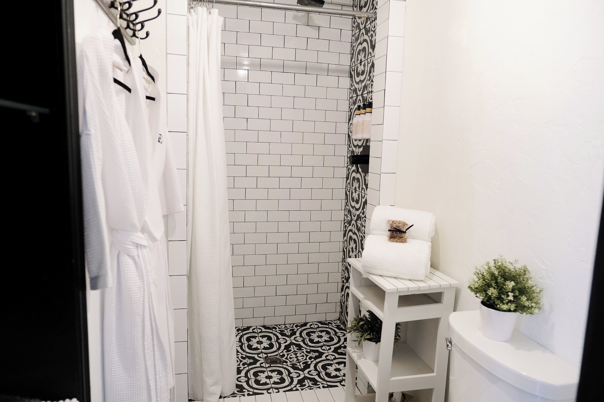 A bathroom with a shower, white tiled walls, a patterned floor, a white sink, and a small potted plant.