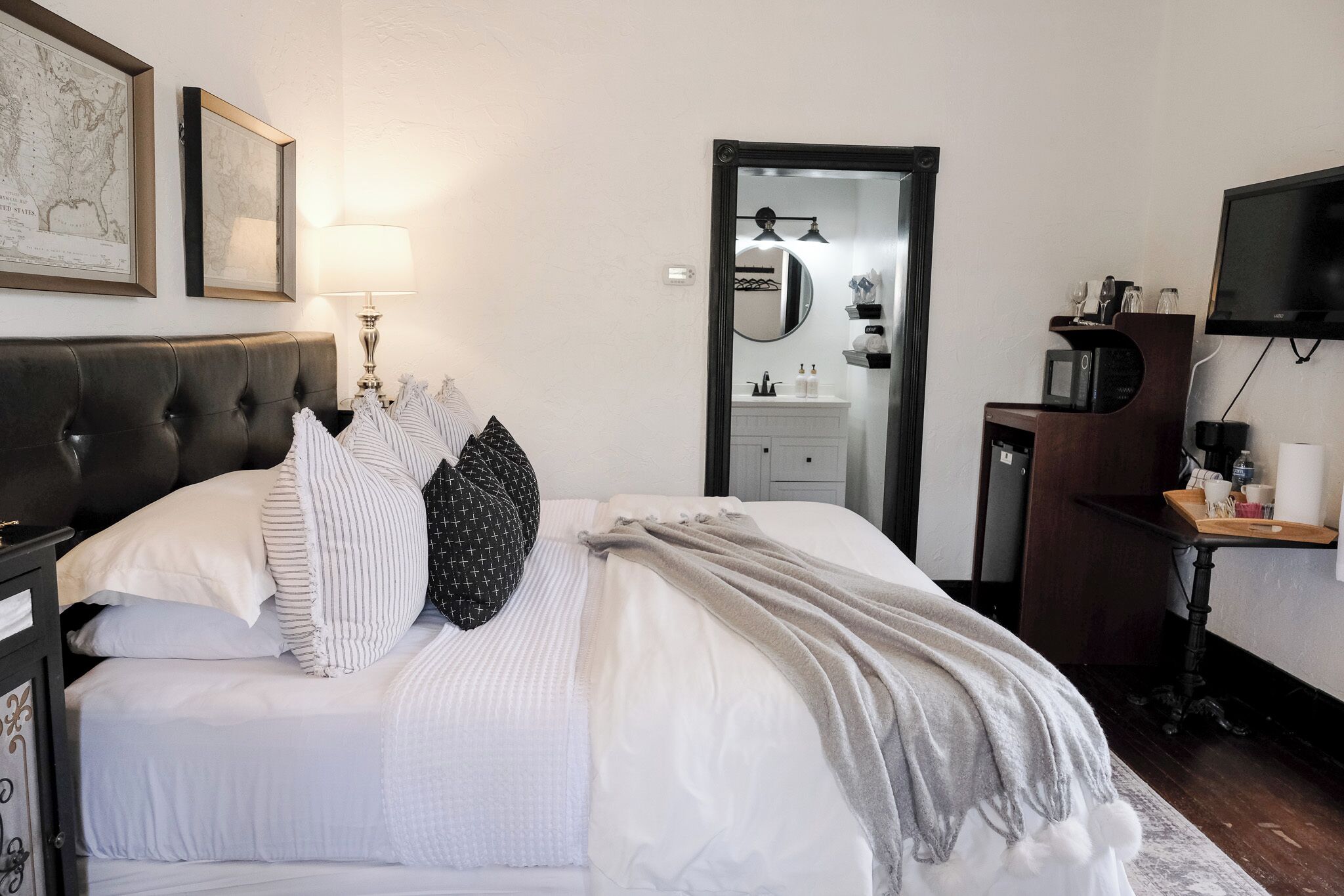 Un lit bien fait, avec du linge de lit blanc et un plaid gris, une table de chevet avec une lampe, et une tête de lit en bois foncé.