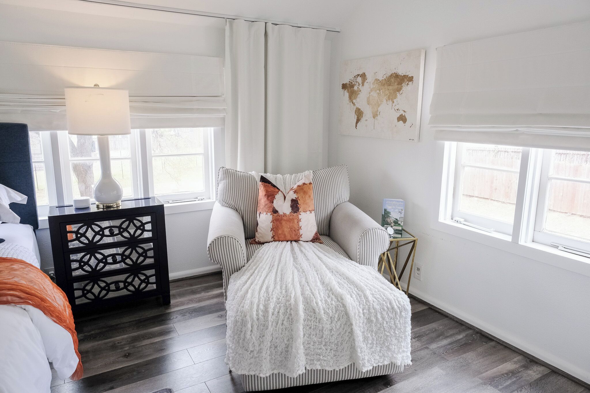 Une chambre à coucher avec un lit, une table de chevet, une lampe, une fenêtre avec des rideaux et une carte du monde accrochée au mur.
