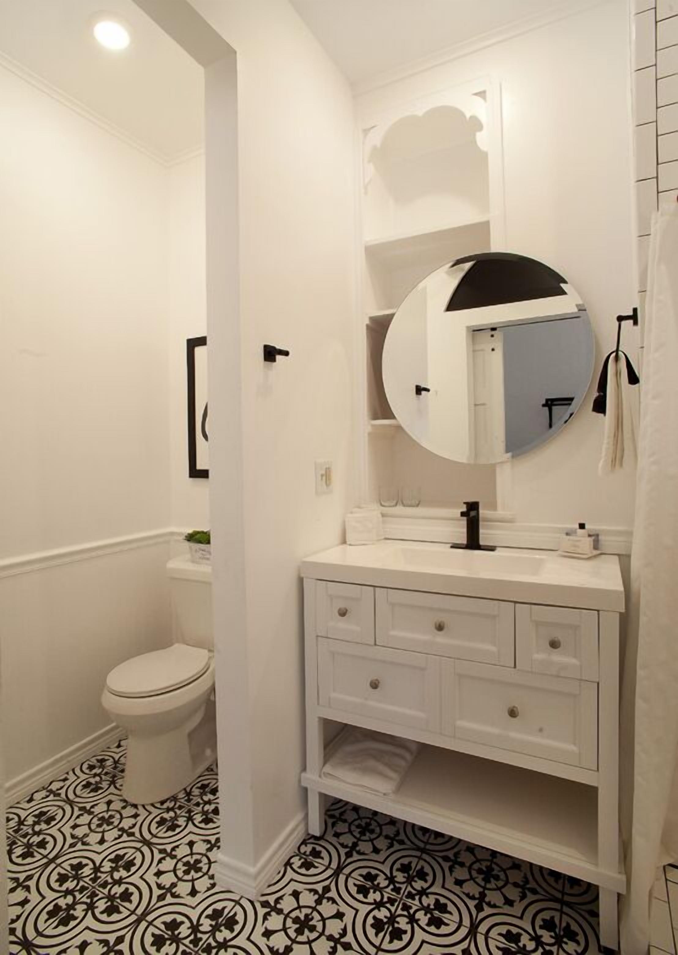 A bathroom with a white vanity, a round mirror, a toilet, and patterned floor tiles.