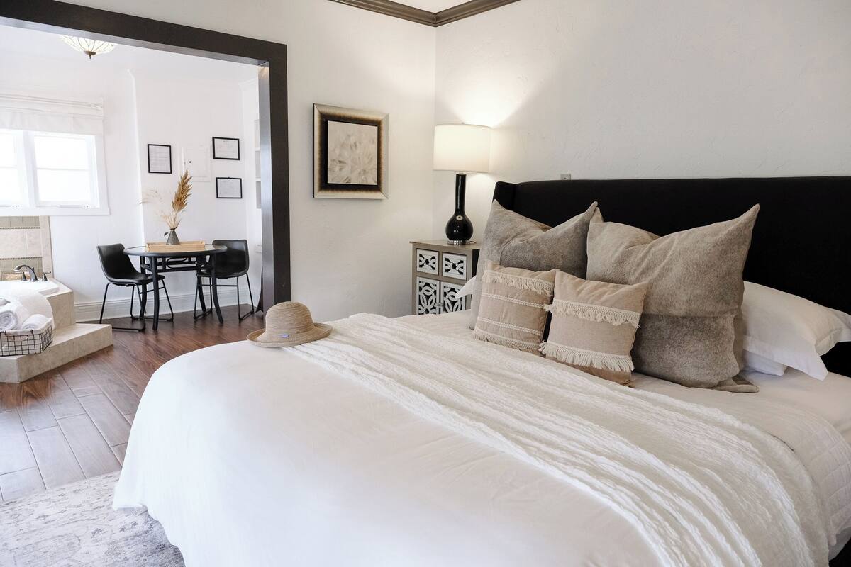 A neatly arranged bedroom with a large bed, bedside table, lamp, and a small desk with chairs.