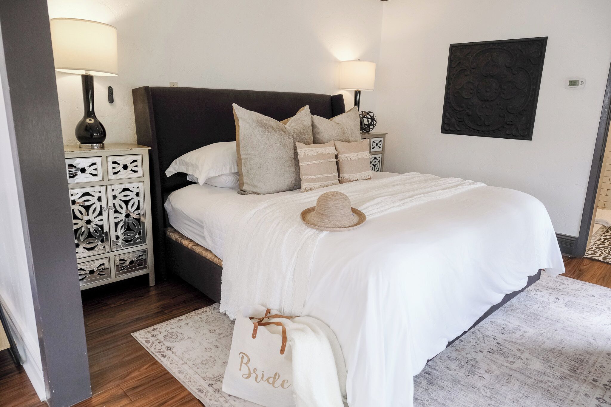 Un lit bien fait, avec une couette blanche, un chapeau décoratif et un sac fourre-tout sur lequel est écrit « Future mariée », posé sur le sol.