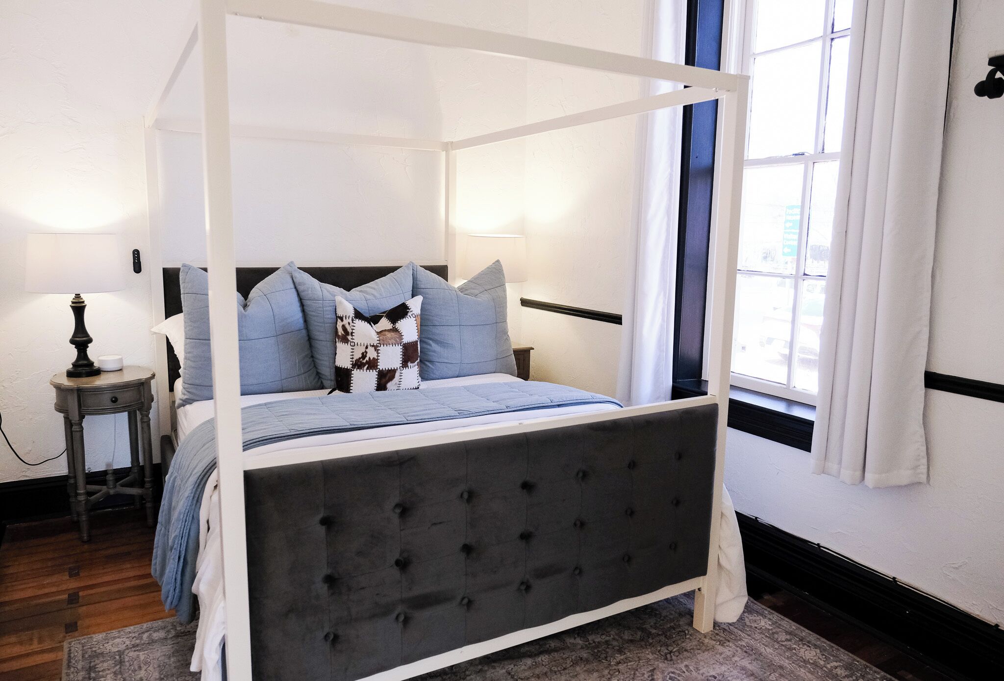 A four-poster bed with a tufted headboard, a bedside table with a lamp, and a view of the outside through a window with curtains.
