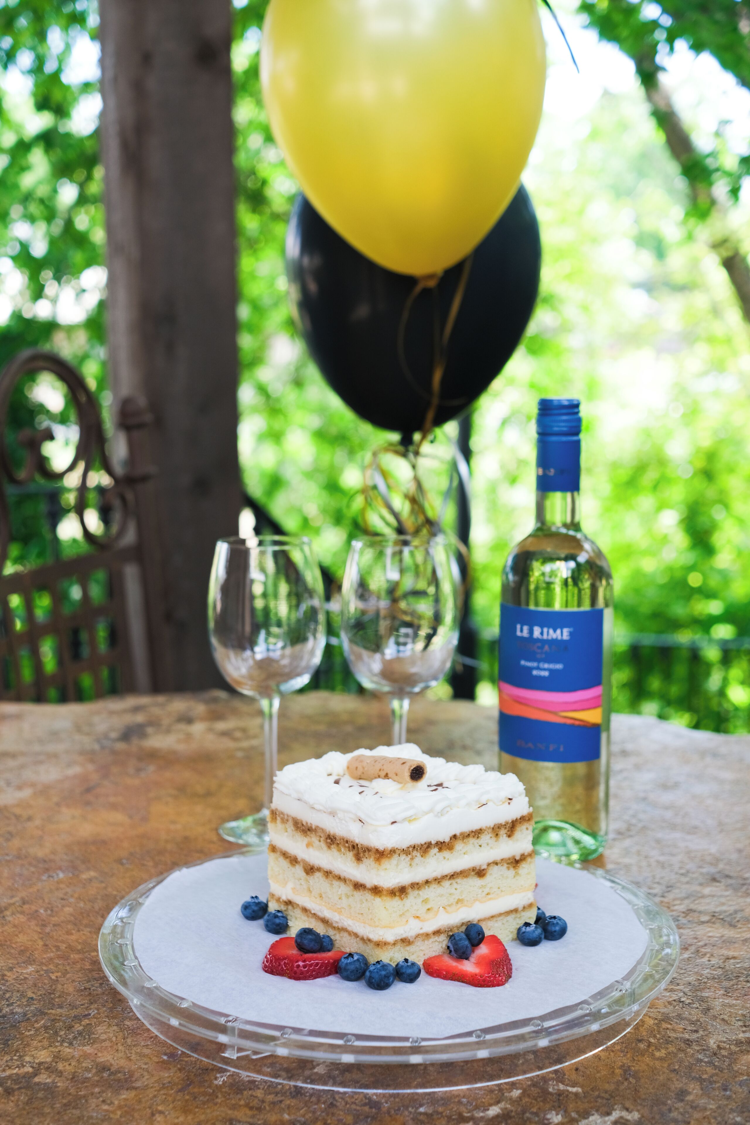 A slice of cake on a glass plate with strawberries and blueberries, two wine glasses, and a bottle of wine on a table outdoors.