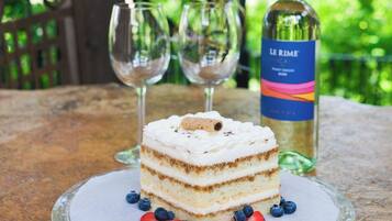 A slice of cake on a glass plate with strawberries and blueberries, two wine glasses, and a bottle of wine on a table outdoors.