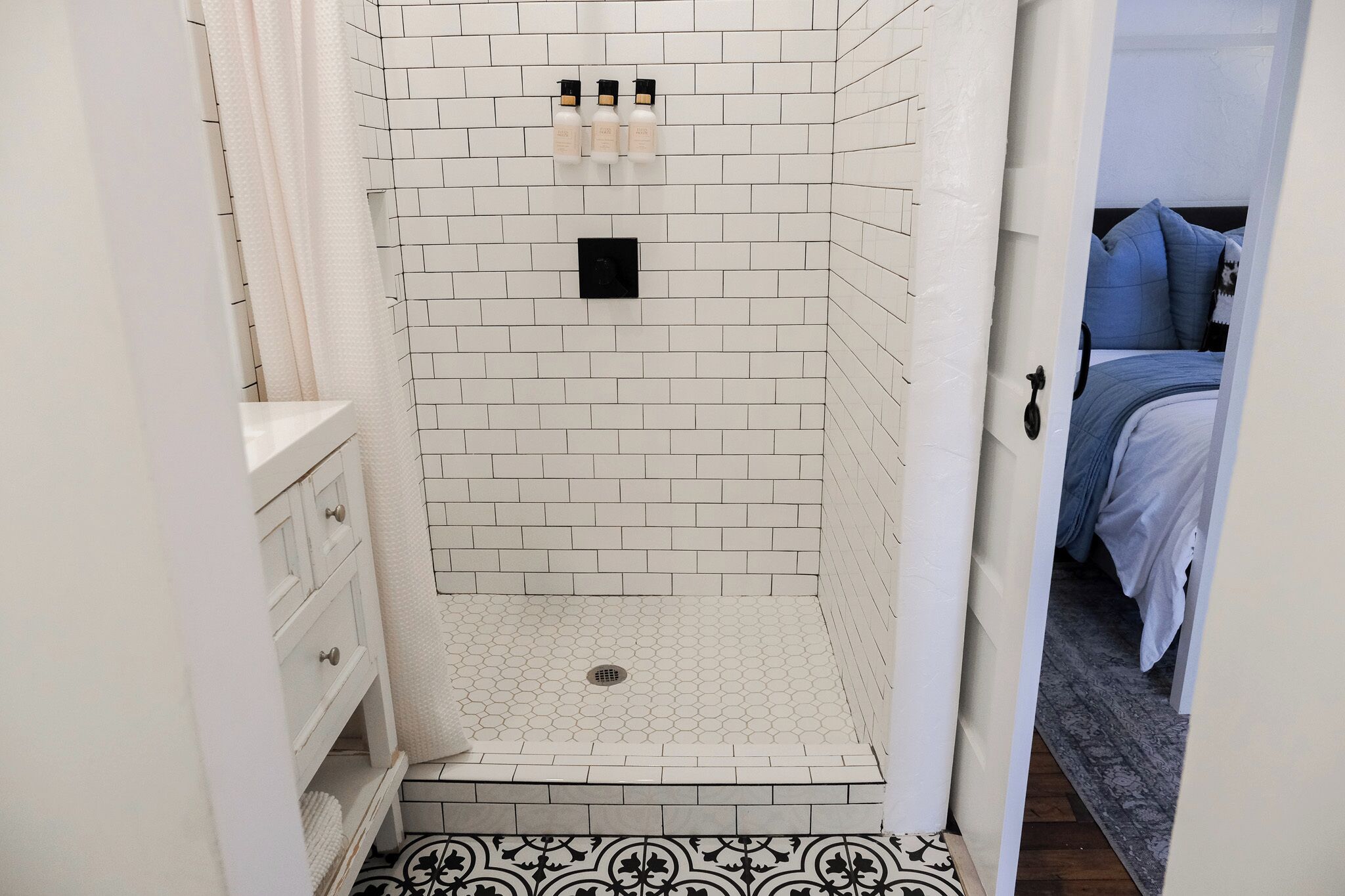 A shower area with white tiled walls, a black control panel, and three bottles of shampoo.