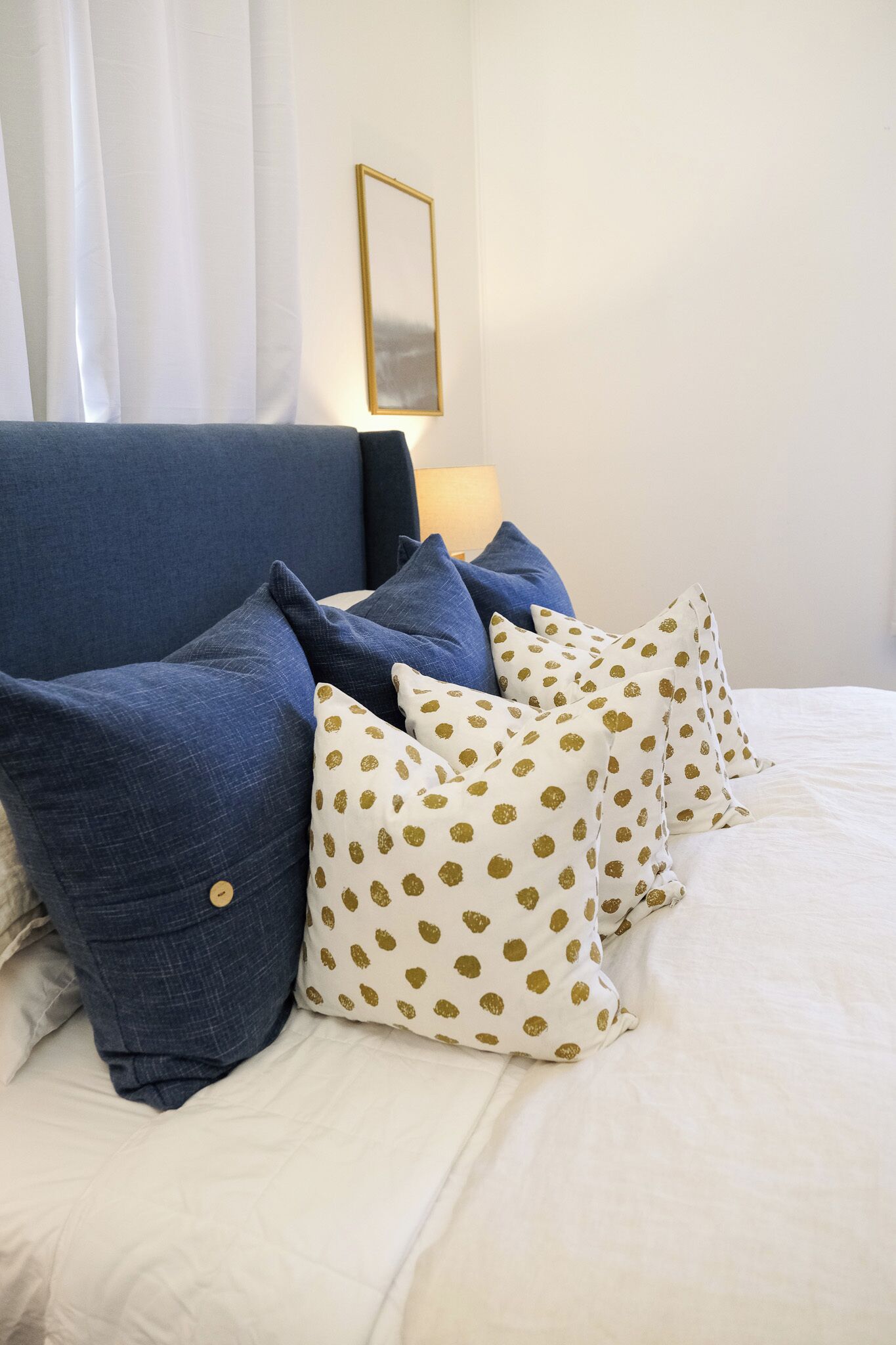 A bed with white bedding and blue and white pillows, a gold-framed mirror on the wall, and white curtains.