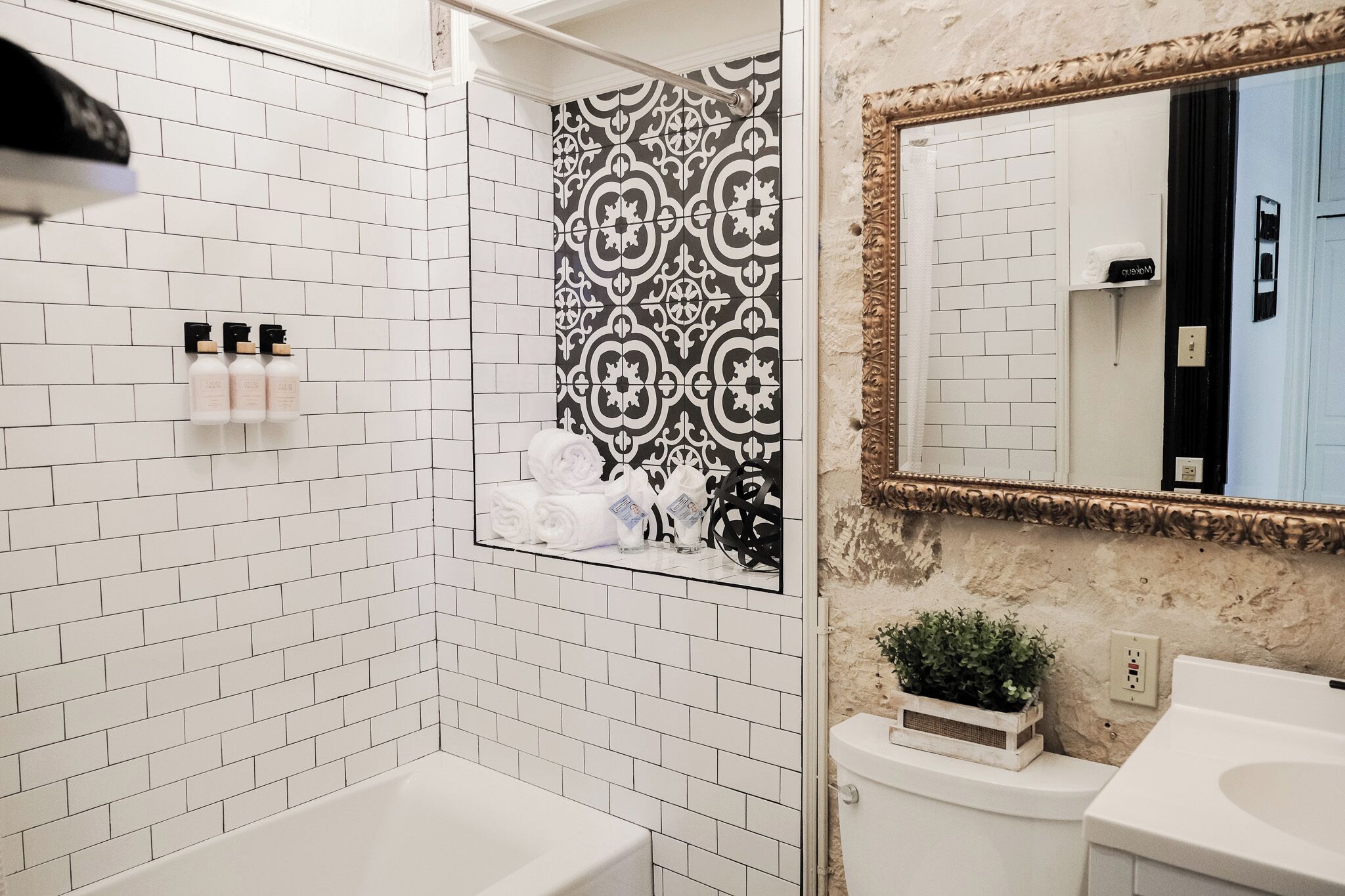 Een badkamer met witte betegelde muren, een douchewand met zwart-wit patroon, een wit wastafelmeubel en een spiegel met gouden lijst.