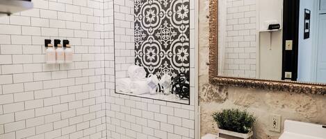 Une salle de bain avec des murs recouverts de carreaux blancs, un mur de douche orné d’un motif noir et blanc, un lavabo blanc et un miroir avec un cadre doré.