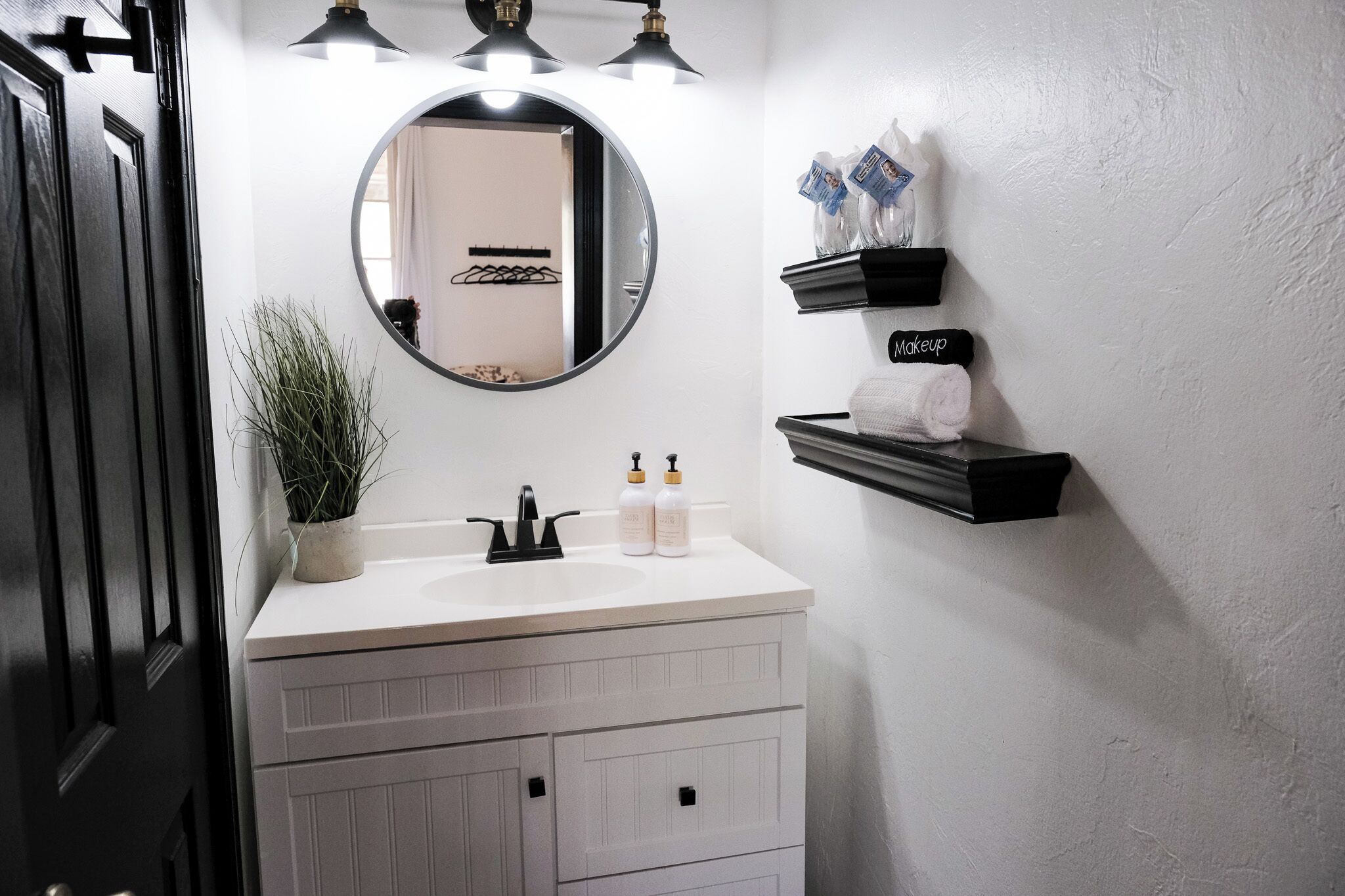 Une salle de bain avec un meuble-lavabo blanc, un robinet noir et un miroir rond. On y trouve deux étagères avec des articles de toilette et une plante en pot.