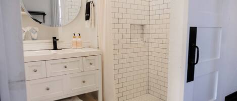 A bathroom with a white vanity, a round mirror, a shower with white tiles, and a black door.