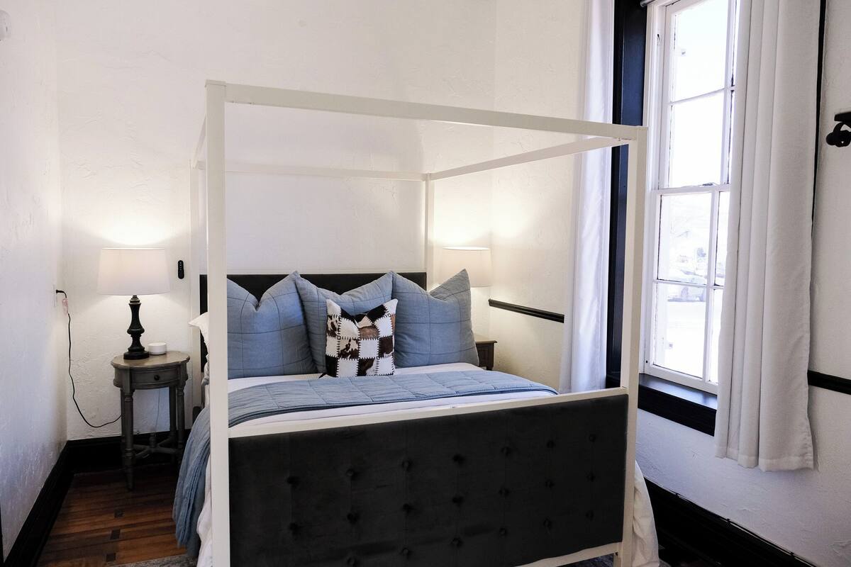 A four-poster bed with a dark headboard, two bedside tables with lamps, and a window with curtains.